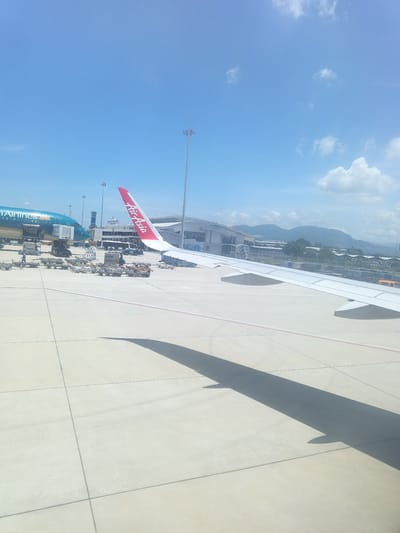 This image captures an airport tarmac scene in Bắc Cam Ranh, Vietnam. In the foreground, a section of a white airplane wing extends across the frame, casting a long shadow on the concrete surface. The wing is attached to an airplane with a bright red tail fin featuring the "Air Asia" logo in white lettering. To the left, another airplane, predominantly teal with "Vietnam Airlines" branding, is parked near a jet bridge. The tarmac is marked with grid lines and a red stripe running parallel to the wing. Various airport equipment, including baggage carts, loaders, and a bus, are visible around the parked aircraft. In the background, airport buildings and terminals are situated, with a mountainous landscape and a clear blue sky with scattered clouds forming the backdrop. The overall scene depicts a typical operational environment at an airport on a sunny day.