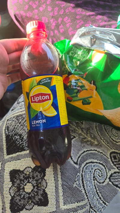 A close-up shot shows a hand holding a Lipton Lemon Ice Tea bottle. The bottle has a red cap and a yellow and blue label that reads "Lipton LEMON ICE TEA LOW IN CALORIES". Behind the bottle, a bag of chips is visible, with green and silver packaging. The subjects are positioned on a patterned blanket. The background is out of focus, but appears to be a person's clothing. There is no indication of the location being in Sakarçäge District, Turkmenistan.