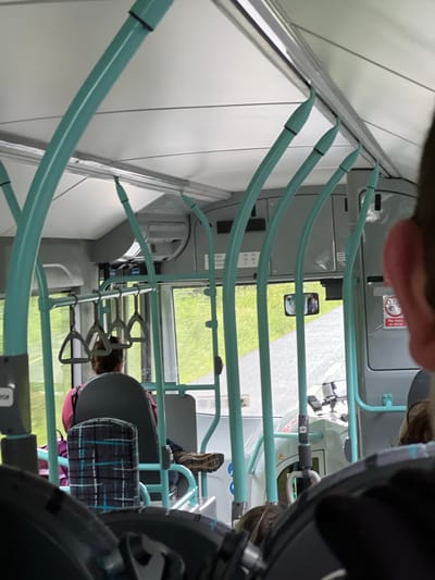 The image shows the interior of a bus on a cloudy day, with a view of green fields and a road through the windows. The bus has teal-colored grab bars and poles. In the foreground, the back of a person's head is visible, wearing dark hair. Further inside the bus, a person wearing a pink top and dark pants sits facing forward. A seat with a plaid pattern is visible in front of this person. The bus appears to be in motion, as indicated by the blurred scenery outside. The image was taken in Tullycullion, Ireland.