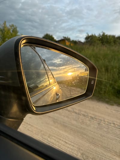 The image is taken from inside a car, focusing on the side-view mirror. The mirror reflects a road surface with tire tracks, suggesting a natural or unpaved road. To the left of the mirror, a blurred landscape with green foliage and trees is visible, under a cloudy sky. The sky appears overcast, with some patches of brighter light filtering through, indicating it is either sunrise or sunset. The reflection in the mirror shows the sun, creating a bright glare and casting warm, golden light on the road and surrounding elements. The overall scene suggests a travel or driving activity during a transitional period of the day, likely in a rural or natural setting within Nizhniye Vyazovye, Russia. No people are visible in the image.