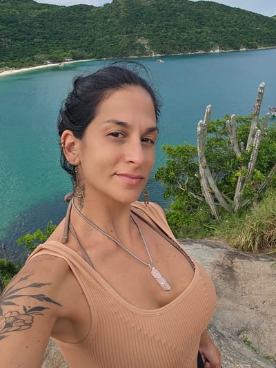 A medium shot captures a woman on a rocky elevated position in Arraial do Cabo, Brazil. The woman, appearing to be in her 30s, has dark hair pulled back and looks directly at the camera with a subtle smile. She wears a light tan ribbed tank top over a brown bikini top. Her visible left ear is adorned with a gold ear cuff and an ornate, silver-toned dangling earring. A silver chain necklace with a wire-wrapped light pink crystal pendant, approximately 5-7 cm in length, rests on her chest. A black ink floral tattoo is visible on her left upper arm.
The background consists of a expansive body of turquoise-blue ocean water. Across the water, lush green, heavily vegetated hills form the distant coastline. A white sand beach with several small structures and boats is visible at the base of these distant hills. To the woman's right, on the rocky terrain, several tall, segmented cacti with prominent spines and green foliage are present. The sky is overcast, indicating diffused lighting conditions. The foreground consists of rough, grey rock.