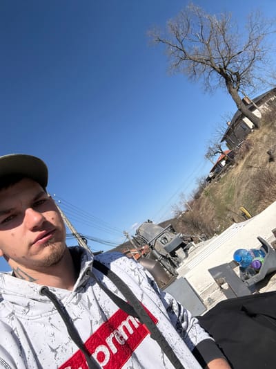 An upward-angled, low-light photograph captures a male individual taking a selfie in Raduil, Bulgaria, under a clear blue sky on a bright day. The subject, a young adult male with light skin, short dark hair, and light stubble, wears an olive-green baseball cap and a white hoodie with a red "Supreme" box logo. Tattoos are visible on the left side of his neck. He looks directly at the camera with a neutral expression.
The background reveals a rural or semi-rural environment. To the left, a utility pole with multiple power lines is present. In the mid-ground, a large, dark-colored piece of heavy machinery, possibly an industrial cement mixer, is angled. The right side of the image features a dry, grassy hillside ascending towards the upper right. Several bare deciduous trees and residential structures, including houses with stone or stucco facades and red-tiled roofs, are visible on the slope. A distinct, slender yellow marker or sign protrudes vertically from the hillside. In the immediate foreground to the right, a white surface holds a gray utility container with at least two plastic water bottles, one with a blue cap and another with a purple cap. A segment of a black object, possibly a bag or fabric, is also visible.