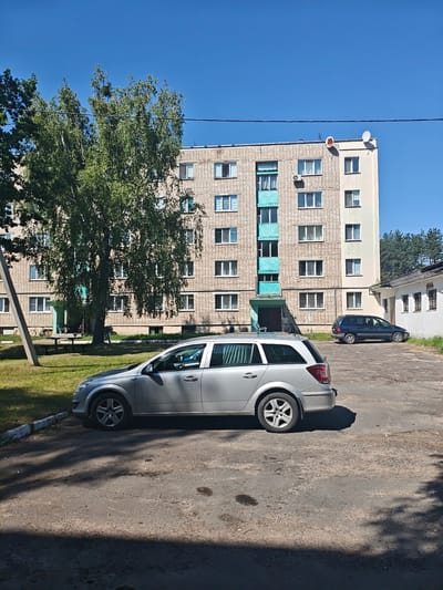 A bright, sunny day is depicted in this outdoor scene, likely in the village of Harochytski, Belarus. A multi-story, brick apartment building with light-colored trim and numerous windows dominates the background. Two satellite dishes are visible on the rooftop. In front of the building, a large, leafy tree casts a shadow over a grassy area that contains a picnic table and benches. A silver station wagon is parked in the foreground on an asphalt surface, with a dark blue minivan parked further back near a white, low-rise building. The ground is textured with asphalt patches and some debris. The sky is a clear, vibrant blue.