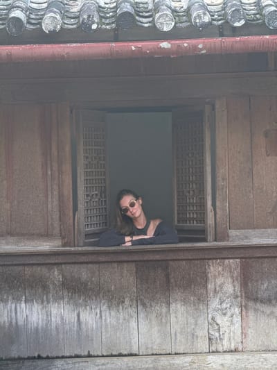 A rotated, medium shot shows a woman peering out of a wooden window in Sà Phìn Commune, Vietnam. The weathered window has a latticed screen. The woman is wearing sunglasses and a dark top. To the left of the window are traditional Vietnamese roof tiles. The wooden wall to the right of the window is aged and weathered.