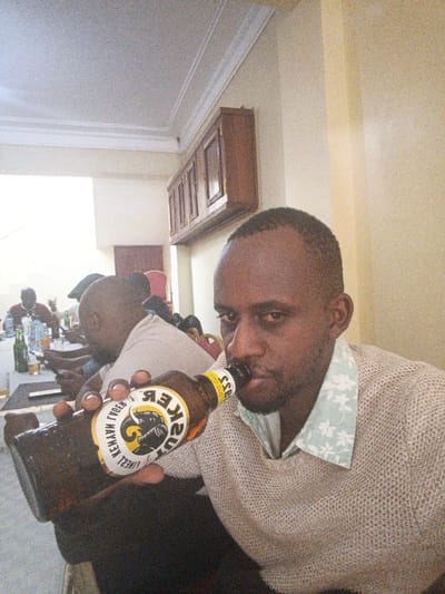 In a warmly lit room, a dark-skinned man drinks from a Tusker beer bottle, held in his left hand. He's wearing a light beige sweater over a floral-patterned shirt. In the background, several other men are seated at a table. More beer bottles, glasses, and phones are visible on the table. A wooden cabinet hangs on the wall behind the table. The room features white walls and a white ceiling with decorative molding.