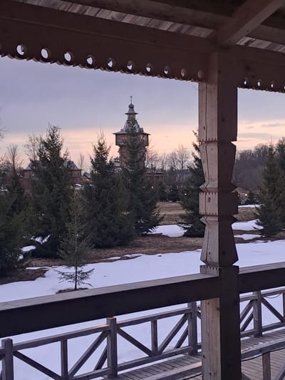 The image shows a view of the Church of the Holy Image of Edessa in Vzglyadnevo, Russia, framed by a wooden structure. The church's spire is visible amongst evergreen trees. Snow is present on the ground. The sky is dusky, suggesting early morning or late afternoon. The wooden structure framing the view appears to be a porch or pergola with decorative cutouts and a supporting post-and-beam design.