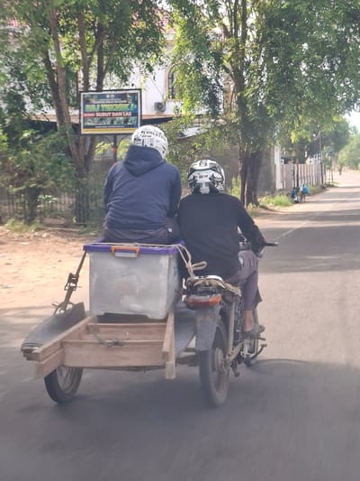 In Meuse, Indonesia, two individuals are riding a motorcycle with a trailer. The person in the front is wearing a blue hoodie and a white helmet with black and silver graphics. The person in the rear is wearing a black long-sleeved shirt and a black helmet with similar graphics. The motorcycle is towing a wooden trailer with a large, gray container with a purple lid. The trailer appears to be constructed from simple wooden planks and has one wheel. The scene is outdoors, with trees and buildings visible in the background. A sign with Indonesian text is mounted on a pole to the left of the road. The road is paved, and there is a dirt or gravel shoulder on the left side. The overall impression is a functional mode of transport for goods or supplies.
