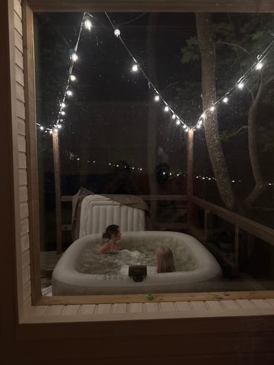 The image shows an outdoor inflatable hot tub at night, illuminated by string lights. Two individuals are visible in the hot tub, submerged in bubbling water. One person, on the left, is facing towards the right with their head and shoulders visible. The other person, on the right, has their back to the camera, with their head and shoulders submerged. The hot tub is positioned on a wooden deck, with trees and foliage visible in the background. A large, white object, possibly a cover or another inflatable item, is leaning against a wooden post behind the hot tub. The scene appears to be in a secluded or wooded area.