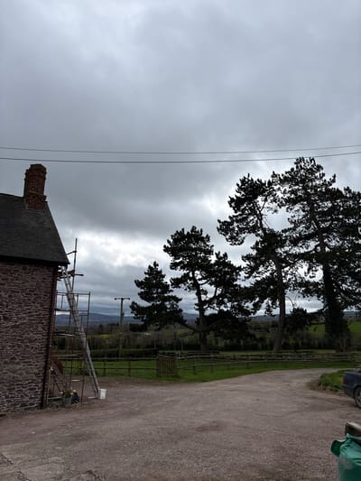 The image depicts a rural scene in Longden, United Kingdom. A stone building with a chimney occupies the foreground, its roof partially covered with scaffolding and a ladder. Overhead power lines are visible against a cloudy sky. In the background, silhouetted trees and a distant landscape with fields, fences, and additional power poles are discernable. A gravel driveway is to the right. A car is partially visible.