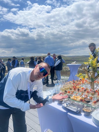 A medium shot shows a man in sunglasses and a white and navy jacket sitting at a table outdoors in Зеленогорское сельское поселение, Ukraine. He has a lanyard with Cyrillic text around his neck. The table behind him holds glasses, snacks, and decorative flowers. Other people are visible in the background, along with a cloudy sky and a landscape.