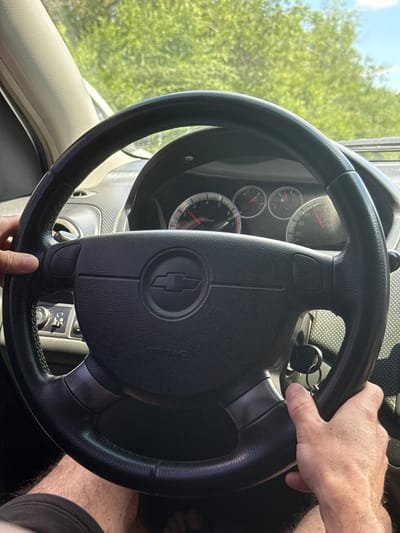 The image shows a close-up view of a person's hands on the steering wheel of a car. The steering wheel has a Chevrolet logo in the center and a horn button on the left side. The car's dashboard, including a speedometer and tachometer, is visible behind the steering wheel. Sunlight streams through the windshield, illuminating the interior of the car and casting shadows. The exterior view through the windshield shows lush green trees and a bright blue sky, suggesting a daytime scene in a rural or suburban setting. The image does not provide any specific information or visual cues to pinpoint the location within the city of Sen'chyne, Ukraine.