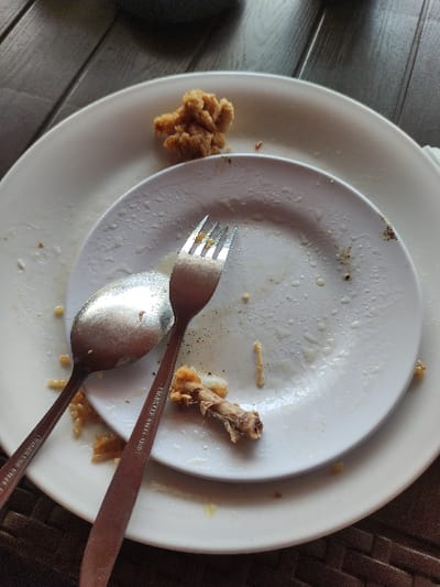 The image shows a white ceramic plate with a small portion of fried chicken and a chicken bone on it. The plate is sitting on top of a larger, white plate with a fork and spoon lying on top. The fork is pointing to the right and the spoon is pointing to the left. The plate is dirty with remnants of food and some black specks. The background shows a wooden surface, likely a table. The image is likely taken inside a restaurant or a dining area, based on the wood grain and the presence of food and utensils. The location of Gambiran, Indonesia, based on coordinates -8.434 latitude and 114.18 longitude, is not directly discernible from the image, and the image offers no contextual information that would allow for a determination of a specific location.