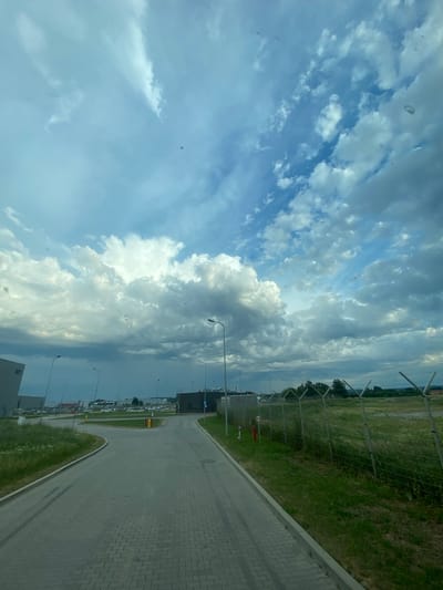 The image depicts an outdoor scene on what appears to be a bright, partially cloudy day, likely in the afternoon given the illumination. The perspective is from inside a vehicle looking outwards, with the image rotated 90 degrees counter-clockwise.
The foreground on the right consists of a paved walkway made of grey interlocking pavers, bordered by a strip of green grass. Beyond the grass is a chain-link fence running horizontally across the frame. Through the fence, a field of green grass extends, with trees visible in the distance. Several lampposts are spaced along the edge of the field, indicating a well-lit area, possibly a public space or industrial zone.
In the mid-ground, a paved road curves away from the viewer, with more lampposts and some buildings. There are parked cars visible in the distance, suggesting a parking lot or an area with activity. A yellow and red fire hydrant is also noticeable near a junction of roads.
The sky dominates the left side of the image, filled with a dramatic display of clouds. Cumulus clouds, both bright white and shadowed grey, are scattered across a pale blue sky, creating a dynamic and textured appearance. There are several small, dark circular marks on what appears to be the glass of the vehicle, one of which is circled in the top left crop.
The scene is set in Niepołomice, Poland. The overall impression is one of an expansive outdoor environment, perhaps an airport, industrial park, or a large recreational area, under a dynamic sky. There is no visible text in the image.