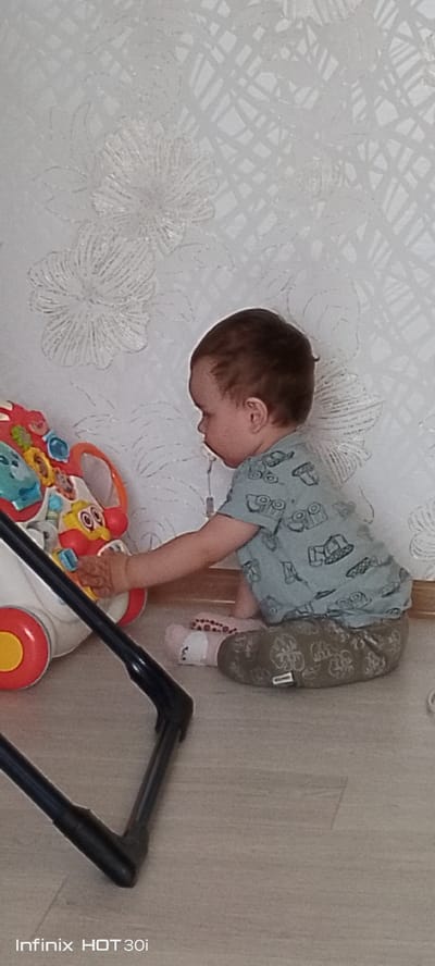 The image shows a young child sitting on the floor next to a walker toy. The child is wearing a light blue t-shirt with a pattern of cars and olive green pants with a leaf pattern. The child is holding a small blue toy. The walker toy is red, white, and yellow with various interactive elements. The background consists of a wall with white textured wallpaper featuring a floral pattern and a light brown floor. The setting appears to be an indoor environment, possibly a home. There is no information in the image that indicates the location in the city of Kedrovy, Russia.
