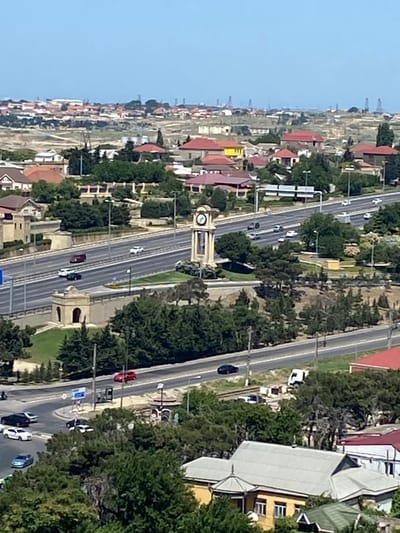 The image shows a high-angle view of a city street in Bakikhanov, Azerbaijan. A multi-lane highway runs through the center of the frame, with several cars and trucks visible on it. To the left of the highway, there is a clock tower and a small, ornate building with an arched entrance. Green trees and manicured lawns surround the highway and buildings. In the background, a cityscape with residential buildings and oil derricks can be seen under a clear blue sky. The overall impression is of a well-maintained urban environment with a mix of modern infrastructure and greenery.