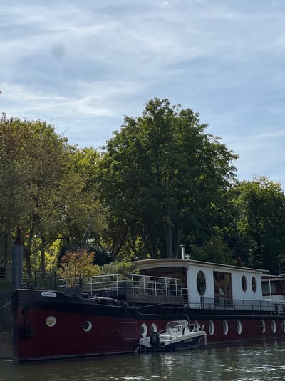 The image displays a static daytime scene in Levallois-Perret, France, featuring a large houseboat moored on a body of water, likely a river. The houseboat, named "L'ARLESIENNE" in white lettering on its bow, has a dark red hull with a black stripe along its upper edge and numerous white circular porthole-style windows. Its white superstructure features larger oval and rectangular windows, a wooden roof overhang, and a white metal railing enclosing a deck furnished with outdoor seating and potted plants. Two silver chimney pipes protrude from the roof. A dark anchor hangs from the houseboat's bow, and a thick mooring rope extends to an unseen point on the bank. A smaller, light-colored motorboat, marked with the registration "HU D5483," is moored directly alongside the houseboat and is equipped with an outboard motor. The water is dark and reflective, showing minor ripples. The background consists of a dense, verdant line of mature trees with varying shades of green foliage, indicating a lush riverbank. The sky is overcast with scattered grey-white clouds, and a dark circular blemish is visible in the upper central sky area. No people or active interactions are visible.