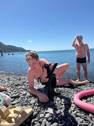 This is a full-body, eye-level shot of two people on a rocky beach. The person in the foreground is a young person, possibly a child, lying on their back on the beach. They are wearing black swim trunks and have wet hair, suggesting they have recently been in the water. They are holding a black snorkel mask with a snorkel attached. Their expression is engaged and playful. The person in the background is an adult male, standing and looking down at the camera. He is also wearing black swim trunks and appears to be wet. The scene is set on a beach with dark, smooth pebbles. The sea is visible behind them, with calm, blue water stretching to the horizon. The sky is a clear, bright blue, indicating a sunny day, likely mid-morning or early afternoon given the strong sunlight. There are some indistinct structures in the water in the background, possibly piers or remnants of older structures. A bright pink pool noodle is visible on the beach in the upper right corner. The overall mood is relaxed and summery. The location is identified as Bol'shoi Utrish, Russia. There is some text visible on a plastic bag near the person in the foreground, but it is not legible.