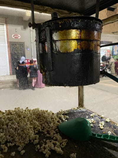 The image displays a close-up, front-facing view of a well-used popcorn machine on a street in Sidi Chahmi, Algeria. The upper cylindrical popping chamber of the machine is heavily coated in dark, burnt oil and caramel residues, with distinct golden-brown streaks. Below this, fresh, white popcorn kernels are scattered across the dark, flat surface of the machine's lower section. A bright green plastic scoop rests on the popcorn to the right.

Through what appears to be a clear, potentially rain-streaked or damp glass enclosure of the popcorn stand, a blurred street scene unfolds in the background. On the left, a building with a partially open white roll-down security door and an ornate dark door is visible. Above the ornate door, a red sign with white Arabic text, reading "حلويات" (sweets/pastries), is affixed. Three individuals stand near this building: an adult female wearing a dark hijab and attire, and two children, one in a dark jacket and light pants, the other partially obscured in what appears to be a pink garment. Further into the blurred background on the right, additional indistinct figures are present, suggesting pedestrian activity. The overall lighting indicates an evening or nighttime setting.