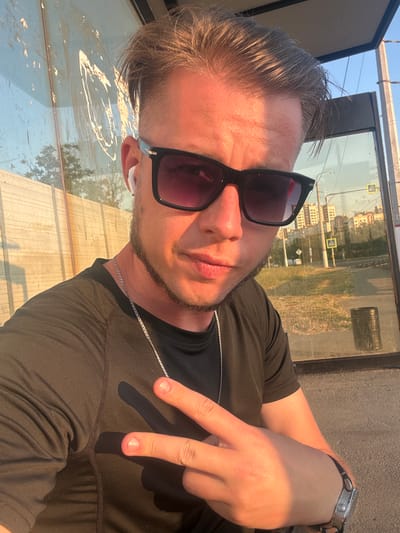 This media file is a selfie taken outdoors. The subject is a person with light skin, short brown hair, and stubble. They are wearing black sunglasses, a black t-shirt, and a silver chain. The person is making a peace sign with their right hand, and a white earbud is visible in their left ear. A watch with a silver band is on their left wrist.

The scene appears to be at a bus stop or similar outdoor waiting area, with a reflective glass panel behind the subject. The reflection shows buildings, trees, and traffic signs, indicating an urban or semi-urban environment. The sun is low in the sky, casting long shadows and warm light, suggesting it is either early morning or late afternoon. The weather appears to be clear and sunny. The location is contextually identified as Мирновское сельское поселение, Ukraine. There is no visible text in the image itself.