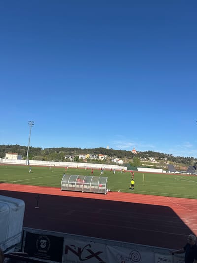The image shows a soccer field in Valpaços, Portugal, with a clear blue sky. The field is bordered by a red running track and advertisements. A group of people, presumably soccer players, are practicing on the field, with some wearing red and others wearing white. A referee in a yellow shirt is also present. A covered bench is located near the track. In the background, there is a hillside with vegetation and some buildings, including what appears to be a church with a prominent spire. A tall light pole is visible near the field.