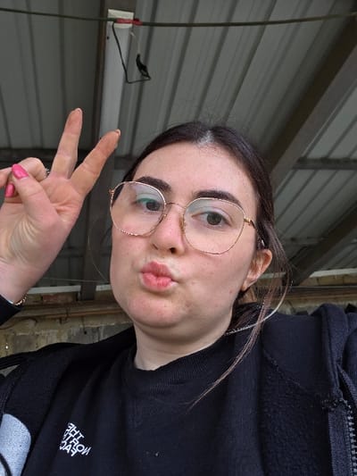 An image taken in Negorci, North Macedonia, depicts a female subject from the chest up, positioned centrally in the frame. The individual has dark hair with some lighter strands visible, and is wearing thin, gold-rimmed eyeglasses. Her lips are pursed in a "kiss face" gesture, and her gaze is directed slightly to the viewer's right. Her left hand is raised, performing a "peace sign" with the index and middle fingers extended; her fingernails are painted pink, and a ring is visible on her ring finger. She is wearing a black long-sleeved shirt, potentially a sweatshirt, with a partial white logo visible on the left side, and a black zippered jacket over it. The background consists of a grey corrugated metal ceiling with structural supports, and a white fixture with black wiring is visible in the upper left quadrant. A light-colored, possibly concrete or brick, wall section is partially visible behind the subject's right shoulder.