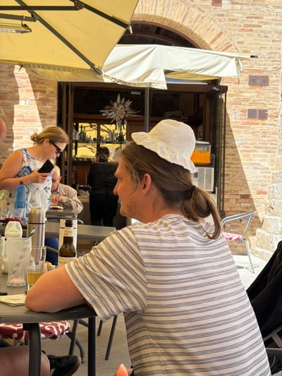 This image captures a vibrant outdoor scene in San Gimignano, Italy, likely at a cafe or restaurant patio on a bright, sunny day. The perspective is unusual, suggesting the photo was taken from above or at an angle, then rotated.

In the foreground, a person with light brown hair tied in a bun is leaning over a dark railing or table. They wear a delicate white bonnet with embroidered patterns and a white and blue striped shirt. Their face is partially visible, looking downwards.

Mid-ground shows a woman seated at a table, wearing sunglasses and a white dress with a blue floral or leaf pattern. She is holding a smartphone and appears to be smiling or laughing. Partially visible behind her is another person seated, wearing a light-colored long-sleeved shirt.

The table holds several items: glasses filled with ice and drinks, a silver thermal flask, a blue plastic bottle, and a bottle with a label clearly showing "GIOLINO". A red and white patterned cloth is draped nearby.

The background features the characteristic brick architecture of San Gimignano, with an arched doorway and windows. One window displays a decorative star-shaped ornament. Two light-colored umbrellas provide shade; one is folded against the building, and another is partially open. Strong shadows suggest mid-day sun. The overall atmosphere is relaxed and social, with people enjoying the pleasant weather and their company.
