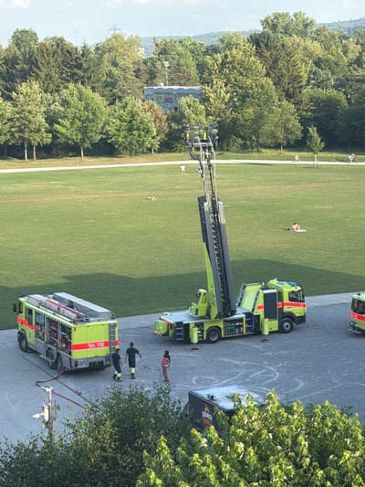 In Glattbrugg, Switzerland, on a bright, sunny day, a scene unfolds in a large, open green park adjacent to a wooded area. Two bright yellow fire trucks are parked on a paved surface. One is a ladder truck with its ladder extended upwards at an angle, and the other is a smaller fire engine. Several individuals are present near the fire engines, including three men in dark clothing and high-visibility vests, and one person in a red outfit. The park's grassy expanse features scattered individuals, some lying down and others in motion, including people on bicycles in the distance. The background reveals a line of trees, beyond which are buildings and a distant power line tower, indicating an urban or semi-urban environment. The sky is clear with a hint of clouds.