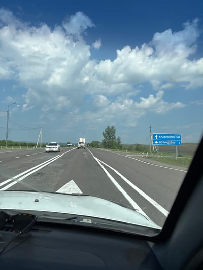 The image shows a highway scene from the perspective of a driver. The road is paved with asphalt and has white lane markings. There are several vehicles on the road, including a white car, a truck, and a white SUV. In the background, there are green trees, fields, and a blue sky with white clouds. A blue road sign indicates that Krasnoyarsk is 343 kilometers away and Polindeika is 3 kilometers away to the left. The weather appears to be clear and sunny. The image does not provide specific information about the city of Borodino, Russia.