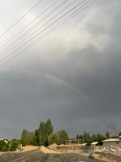 The image shows a landscape scene under a cloudy sky with a faint rainbow visible. In the foreground on the left, several parallel wires run diagonally across the frame. The right side of the image features a sloped dirt path adjacent to a constructed area. This constructed area includes concrete walls, wooden structures, and buildings with white roofs. Beyond the structures, a line of tall, slender trees stands out against the darker sky, with mountains visible in the far background. The weather appears overcast, possibly after rain, given the presence of the rainbow. The time of day is likely late afternoon or early evening, as suggested by the lighting. The scene appears to be rural or semi-rural, consistent with the provided location context of Bujum, Kyrgyzstan. There is no visible text in the image.