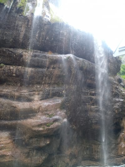 The image depicts a waterfall cascading down a rough, textured rock face. The rock appears to be composed of layers and crevices, with patches of moss growing in some areas. Water streams down in various thicknesses, creating a shimmering effect. Sunlight is visible in the upper right portion of the image, creating a bright glare. A portion of a white, modern structure, possibly a bridge or walkway, is visible at the very top right edge of the frame. The environment appears to be natural, with greenery visible in the distance. The specific location in Khushto-Syrt, Russia, is not visually identifiable from the image content alone. There are no people or animals visible in the frame.
