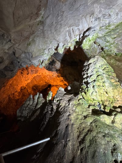 The image shows the interior of a cave, likely the Sveta Petka cave located in North Macedonia. The cave features natural rock formations, including stalactites and stalagmites, some of which are illuminated with an orange light, creating a dramatic contrast with the darker areas of the cave. Greenish moss or algae is visible on some of the rock surfaces, particularly on a large, textured formation in the foreground. A metal handrail or railing runs diagonally across the lower left portion of the image, suggesting it is a tourist-accessible site. The overall environment is dark and cavernous, with the lighting highlighting the geological features.