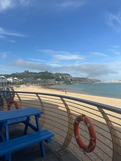 This is a photograph of the seafront in Dover, United Kingdom, likely taken from a raised promenade or balcony overlooking the beach and harbor. In the foreground, a bright blue picnic table and bench are visible, with a metallic railing and a life preserver hanging on it. The beach is covered in light-colored pebbles, and beyond it is the sea. On the horizon, the white cliffs of Dover are prominent, topped by Dover Castle. The harbor area features several large structures, including what appears to be a cruise ship. The sky is a clear blue with scattered white clouds. A few people can be seen walking on the beach and along the promenade. The overall impression is of a bright, sunny day in a coastal town.