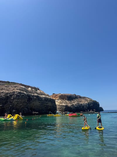 The image captures a vibrant scene at an outdoor water park in Mellieħa, Malta. The crystal-clear turquoise water dominates the foreground, reflecting the bright blue sky. Several inflatable water toys, including yellow slides and red bumper boats, are scattered across the water, suggesting a lively recreational area. In the middle ground, two individuals are paddleboarding, each on a yellow board and wearing life vests, actively engaging in the water activity. Their reflections are visible on the calm surface of the sea.
The background features a dramatic rocky cliff face, characteristic of the Maltese coastline, with a clear demarcation between the dark, rugged lower sections and the lighter, more weathered upper portions. A few people are visible on the beach or near the cliffs, enjoying the sunny day. The overall impression is one of a picturesque and enjoyable summer day at a popular coastal attraction in Mellieħa.