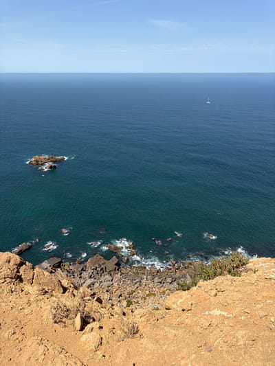 This is an aerial, vertical shot of a rocky coastline meeting the deep blue sea under a clear, bright sky. The scene is split vertically, with the left side dominated by a vast expanse of water transitioning into a pale blue sky on the far left. A small white sailboat is visible on the horizon in this oceanic expanse. The right side of the image features a rugged, sun-drenched cliff face with a textured, sandy-brown surface. Lush green vegetation clings to the upper edge of the cliff, cascading down towards the water. The base of the cliff is strewn with large, dark rocks, where the waves crash against them, creating white foam. The sun appears to be high in the sky, suggesting it is daytime, and the weather is clear and fair. The overall impression is one of a dramatic natural landscape, likely a popular viewpoint overlooking the Atlantic Ocean in Colares, Portugal. No text is visible in the image.
