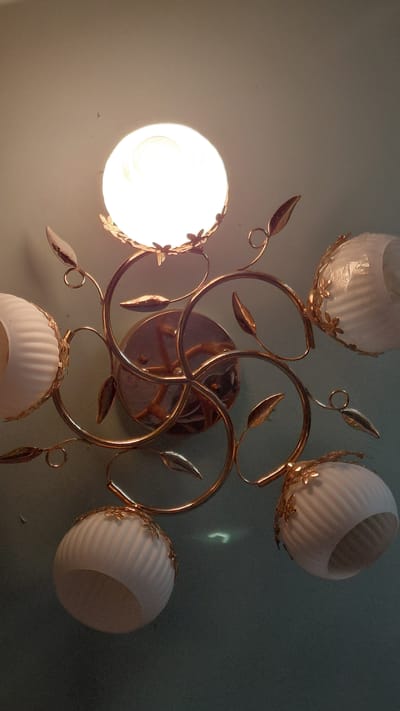 The image shows a ceiling light fixture with a central globe lamp and four smaller globe-shaped shades. The fixture has a decorative design with swirling metallic branches and leaf-like accents. The shades are made of white, ribbed glass, and the central globe is illuminated, casting a bright light. The background appears to be a wall or ceiling painted a pale green or blue. There is no indication of the city of Ak-Terek, Kyrgyzstan, in the image.