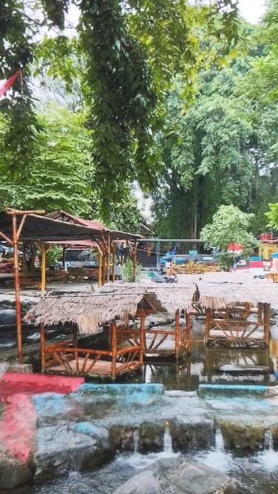 This image depicts a recreational area in Palangka Raya, Indonesia, characterized by a naturalistic setting combined with man-made structures. The foreground features a multi-tiered water flow system with small waterfalls, constructed from grey stone or concrete, parts of which are painted red and light blue. Two open-sided, thatched-roof huts, constructed from wooden or bamboo poles, are situated over the shallow water, appearing to be seating or dining platforms. To the left, a larger, open-air structure with a dark-colored roof supported by bamboo poles is visible on solid ground. The background is dominated by dense, lush green tropical foliage, with taller trees forming a canopy. Further back, a cleared area contains various brightly colored objects, including blue and yellow structures, and red and white striped elements, possibly tents or market stalls. A section of a vehicle, potentially a bus or truck, is partially visible behind the larger covered structure. No individuals are present within the frame. The scene appears to be an unoccupied leisure or dining establishment.