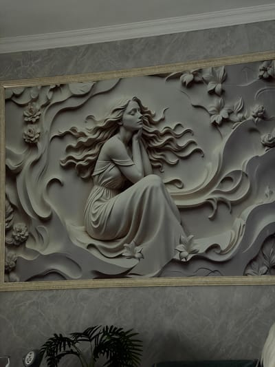 The image displays a close-up of a relief sculpture mounted on a wall. The sculpture features a woman with flowing hair, seated and leaning on her hands. She is depicted in a classical style, wearing a draped dress. Surrounding her are stylized floral and wave-like elements. The sculpture is housed within a decorative frame, and the wall it's mounted on has a marble-like texture. The scene is illuminated by ambient lighting, casting subtle shadows that highlight the relief's contours. The photograph does not provide information about the city of Dzhalgan, Russia, nor does it depict any discernible environmental context beyond the artwork and its immediate surroundings.