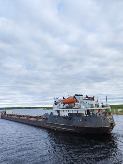The image shows a cargo barge on a body of water. The barge is grey and laden with cargo, with a raised section at the rear containing the ship's bridge and safety equipment. The word "ШАЛА" (Shala) is visible on the stern, along with other inscriptions in Russian. The water is dark with ripples, and the sky is overcast with numerous clouds. A strip of land, perhaps a riverbank or shoreline, is visible in the background. The barge appears to be in motion. The city is not mentioned in the image or in any way visible in the image.