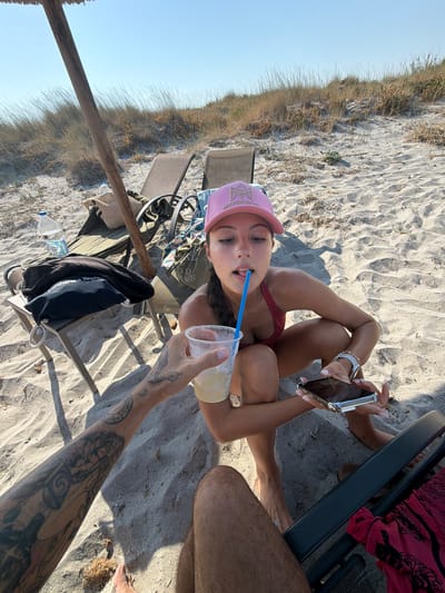 A person wearing a pink cap and a red bikini top is lying on the sand at a beach. They are holding a phone and a drink with a blue straw. Another person's legs and arm with tattoos are visible. The scene is set during the day with clear sky, suggesting good weather. The location is Moudros Municipal Unit, Greece, indicated by the beach setting. The person appears relaxed and is enjoying a drink on a sunny day.
