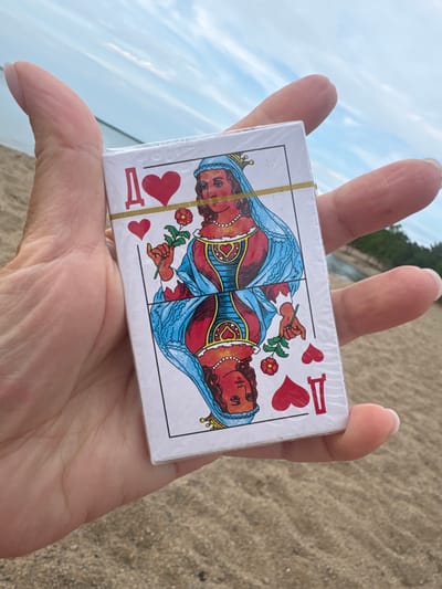 A close-up shot shows a person's hand holding a deck of playing cards. The cards are wrapped in clear plastic and feature the image of a queen on the face. The queen is depicted with a blue headdress and is holding a rose. Red hearts and the letter "Q" are also visible on the card. The background is blurred, showing a sandy beach and a body of water under a cloudy sky. The overall lighting suggests it is daytime, possibly late afternoon or early morning, given the soft light. The scene evokes a sense of relaxation and leisure, possibly a vacation or a casual outing at the beach. The person's fingers are gently holding the card deck, suggesting careful handling. The sandy texture of the beach and the water in the background point to a coastal or lakeside setting.