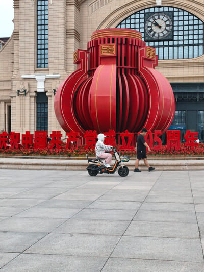 In front of a light-colored brick building with arched windows and a large clock, a giant red lantern sculpture is displayed. Red Chinese characters are visible in front of the sculpture. A person rides a small electric scooter on a paved plaza, and another person walks to the right of the scooter. The scene appears to be a public square in Harbin, China.