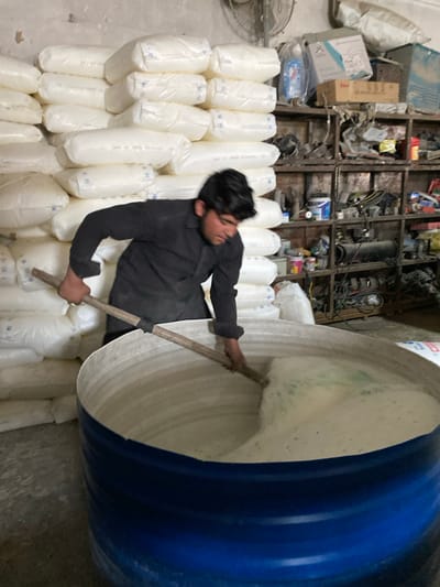 In an indoor industrial setting within Daman, Afghanistan, a young adult male is actively stirring a light-colored, frothy substance contained within a large, blue cylindrical vat. The male, positioned centrally, wears a dark long-sleeved shirt and uses a long wooden pole to agitate the mixture. To the left, a tall stack of numerous white, plastic-wrapped sacks or bags is visible against a wall. To the right, a cluttered shelving unit holds various objects including boxes, a blue bottle, and other miscellaneous items. A ceiling fan is partially visible in the upper right background. The room's walls appear light-colored and somewhat unfinished.