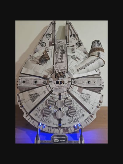 A detailed model of the Millennium Falcon spaceship from Star Wars is displayed indoors. The model is primarily light gray with intricate detailing and appears to be made of wood or plastic. It rests on a dark gray base with blue LED lighting illuminating the lower portion of the model and casting a glow on the surface it is placed upon, which appears to be a wooden desk or table. The background is a plain, light-colored wall. The base of the model has the text "Millennium Falcon™", "STAR WARS", and "UGEARS" printed on it. There are no people or animals visible in the image. The setting is a still, indoor environment. The image does not provide any information or visual cues about a specific location in the city of Norcia, Italy.