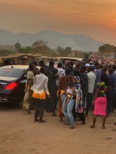 A large crowd surrounds a black car on a dirt road, near a mountain range. The crowd is a mix of ages and attire. Some people are wearing colorful dresses, jeans, and patterned shirts. A woman in the crowd holds a baby. In the background, there is a sign, some trees, and the sky. The image has warm tones, possibly from the light of the setting or rising sun. The overall atmosphere seems to be busy and potentially celebratory.