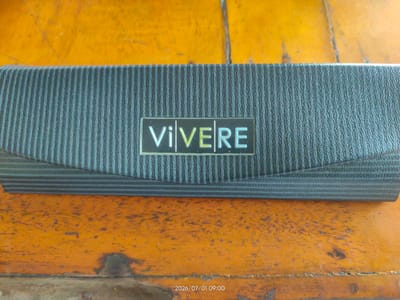 A rectangular, dark gray, textured case is centered horizontally in the image. The case has a subtle wave pattern running horizontally across its surface and a metallic emblem with the word "VIVERE" in white and light green letters. The case rests on a weathered, orange-colored surface that appears to be wood, with visible grain and dark marks. The background is a solid orange surface. A timestamp "2026/07/01 09:00" is visible in the bottom right corner of the image. The image was taken in Geulumpangpayong, Indonesia.