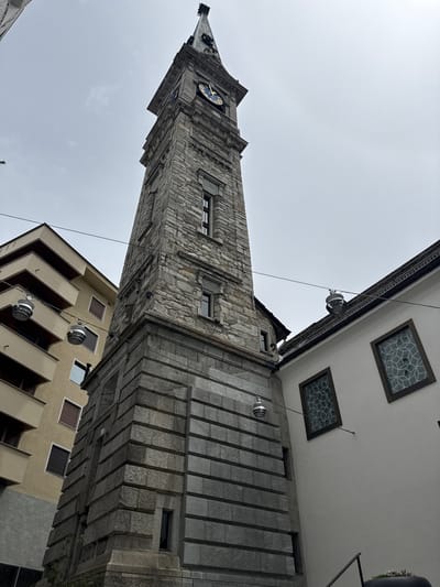 The image shows the clock tower of the former Falck steelworks in Sesto San Giovanni, Italy, which has been converted into a museum and cultural center. The tower, made of stone, rises to the right of the frame, with a clock face visible near the top. To the left of the tower is a modern apartment building with multiple balconies. To the right of the tower is a white building with decorative windows. Several pendant lights hang from wires strung across the scene. The sky is overcast.