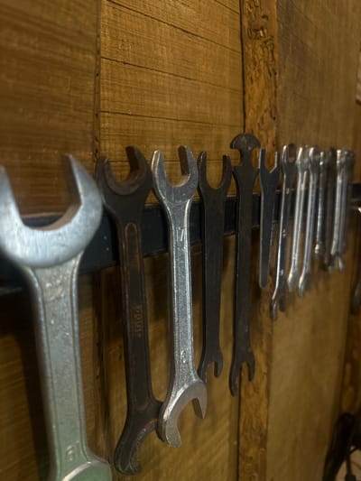 A close-up, eye-level shot shows a collection of wrenches hanging on a rack against a wooden wall. The wrenches are arranged in a line, with the largest one on the far left. The wall is made of horizontal wooden planks, with a vertical wooden beam on the right. The lighting is dim, with a warm tone, suggesting an indoor setting. The scene appears to be a workshop or garage. This image was taken in Lapės, Lithuania.
