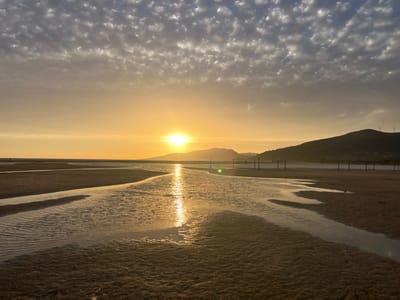 This photograph captures a serene sunset over a sandy beach in Tarifa, Spain. The sun, a bright orb, is low on the horizon, casting a warm, golden glow across the scene. The sky is filled with scattered clouds, which are illuminated by the fading sunlight, creating a textured and dramatic effect.
In the foreground, the wet sand of the beach is marked by shallow channels of water, reflecting the golden hues of the sky. These channels create a sense of depth and movement within the image. Further out, the sea stretches towards distant, silhouetted hills, which are topped with wind turbines, hinting at the area's commitment to renewable energy.
The overall atmosphere is peaceful and tranquil, evoking the feeling of the end of a day. There are a few indistinct figures visible in the distance, adding a subtle human element to the expansive landscape. The image is a testament to the natural beauty of Tarifa, a place known for its stunning beaches and breathtaking sunsets.