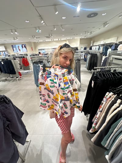 A woman is standing in a retail clothing store, holding up two pairs of patterned shorts. The shorts feature cartoon characters and text. The woman is wearing a red and white checkered skirt, pink sandals, and sunglasses pushed up onto her head. She has blonde hair in a ponytail. The store appears to be well-lit with ceiling lights and displays of clothing on racks and shelves. The flooring is light-colored tile. The background shows aisles of clothing racks stocked with various garments, including jeans, shirts, and other shorts. A sign displaying prices and merchandise information is visible on the shorts the woman is holding. The store is located in Polígono Industrial De Quart De Poblet, Spain, as indicated by the price tag format and the signage visible in the store.