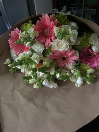 A bouquet of pink and white flowers, including gerbera daisies and snapdragons, is wrapped in brown paper. The flowers are arranged in a circular shape, with the stems facing inward. The bouquet is located in Río Oro, Costa Rica (latitude: 9.945, longitude: -84.201). The bouquet is resting on a dark surface. There are no people in the photo.